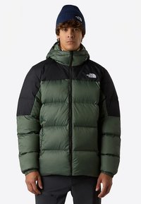 Vadderad jacka med en olivgrön kropp och svarta axlar. Har en dragkedja framtill, huva och en logotyp på vänster bröst.