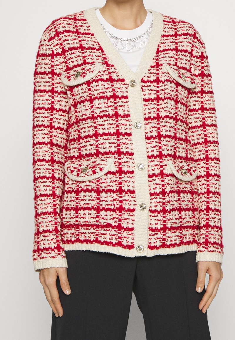 Cardigan en maille à carreaux rouges et blancs avec bordure crème, boutons décoratifs, quatre poches à rabat, porté sur un haut blanc et un pantalon noir.