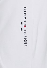 White cotton fabric features "TOMMY HILFIGER" embroidered in black with a red and blue accent, and "EST. 1985" below in black thread.