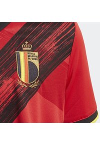 Maglia sportiva rossa con una striscia diagonale nera, stemma del Belgio ricamato con corona dorata e alloro, e dettagli in tessuto testurizzato.