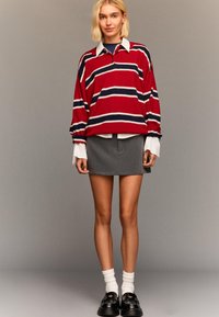 Rdeče in temno modre črtaste velike pulover s zadrgo, v kombinaciji s sivo mini krilom. Bele manšete srajce so vidne. Črni loafers in bele nogavice.