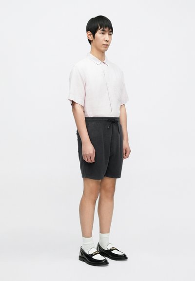 Chemise rose pâle à manches courtes avec boutons, short gris foncé avec cordon de serrage, chaussettes blanches, mocassins noirs et blancs avec accents dorés.