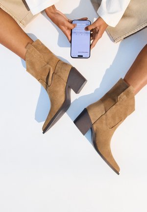 Stivaletti alla caviglia in suede beige con tacco a blocco, caratterizzati da una silhouette curvata e dettagli di cucitura sottili, esposti insieme a uno smartphone.