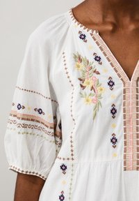 Vit bomullsklänning med puffärmar, prydd med färgglad broderi och detaljerad sömnad. Framträdande blom- och geometriska mönster förhöjer designen.