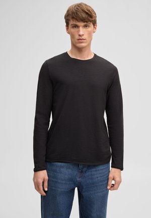 Jonge man staand gekleed in een effen zwarte lange mouwen shirt en blauwe spijkerbroek tegen een neutrale grijze achtergrond