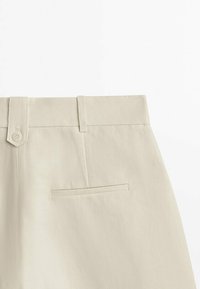 Pantalon beige clair taille haute avec passants de ceinture, patte boutonnée à la taille et poche fendue horizontale à l’arrière côté droit.