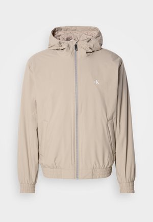 Hellbeige Windbreaker-Jacke mit Kapuze, elastischen Bündchen und Saum, ausgestattet mit einem Frontreißverschluss und einem kleinen Logo auf der Brust. Glatte Textur.