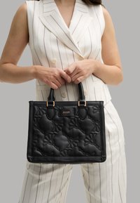 Borsa a mano nera trapuntata con motivo floreale in rilievo e dettagli dorati. Presenta manici doppi e un'etichetta con logo. Texture liscia.