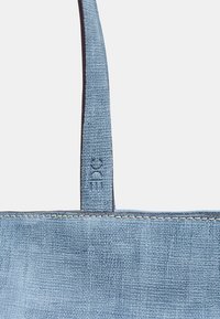 Sac fourre-tout bleu clair en tissu texturé, avec un bord cousu et une sangle de marque ornée d'un logo en relief.