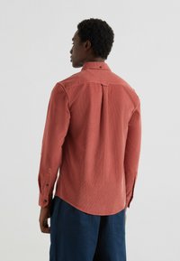 Camicia di velluto a coste rossa con maniche lunghe, polsini con bottone e una piega sul retro. Presenta un colletto tradizionale e una texture liscia.
