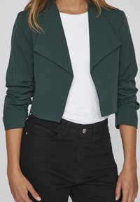 Afskåret grøn blazer med sjalkrave, elastiske ærmer, parret med en hvid top og sorte højtaljede jeans. Glat tekstur.