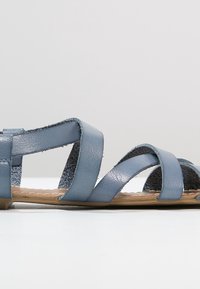 Sandales en cuir bleu avec des brides croisées, une semelle plate et une semelle intérieure texturée. Le design est minimaliste et ouvert.