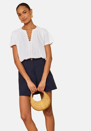 Kvinde iført hvid bluse med bølgede V-hals og marineblå shorts, der holder en rund flettet taske mod en ensfarvet baggrund.
