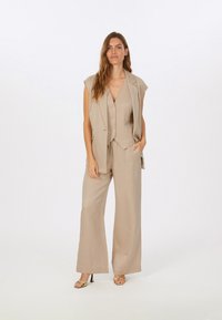 Beige maßgeschneideter Dreiteiler mit einem ärmellosen Blazer, einer Weste mit Knöpfen und weit geschnittenen Hosen aus einem glatten Stoff.