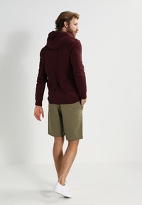 Bordeauxkleurige hoodie met ribbels aan de manchetten en zoom, gecombineerd met olijfgroene shorts. Model staat met de rug naar de camera en draagt witte sneakers.