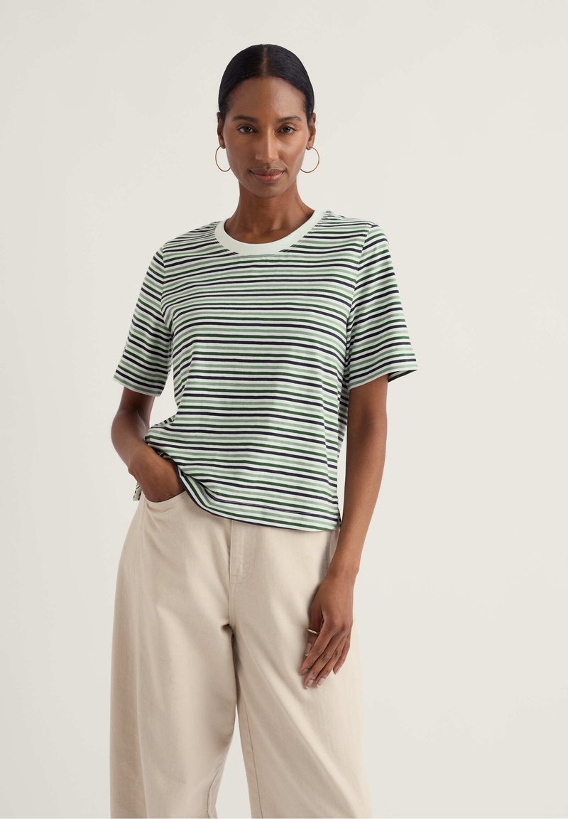 Žena v krátkém rukávu s bílým, černým a zeleným proužkovaným tričkem, částečně zastrčeným do béžových širokých kalhot, stojící s jednou rukou v kapse.