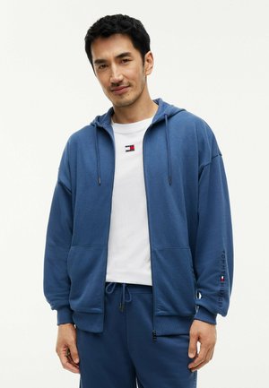 Man draagt een blauwe hoodie met rits en bijpassende broek met een wit T-shirt, staand tegen een effen lichte achtergrond.