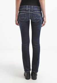 Kvinna som bär mörkblå smalpassande jeans och svarta skor, stående med ryggen mot en vit bakgrund.