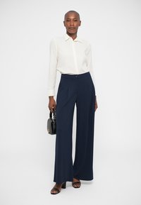 Balta ilga rankovė marškiniai su mygtukų uždarymu, derinami su tamsiai mėlynomis plačiomis kelnėmis. Modelis laiko mažą juodą rankinę ir užsideda juodus šlepetes.