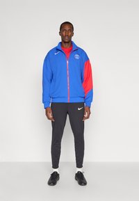 Férfi áll kék és piros Paris Saint-Germain dzsekiben, fekete Nike nadrágban, fehér zokniban és fekete sportcipőben egy sima háttér előtt.