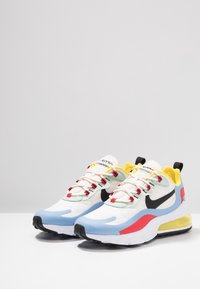 Paire de baskets blanches Nike Air Max avec un swoosh noir, des accents bleus, rouges et jaunes, sur une surface blanche devant un fond gris clair.