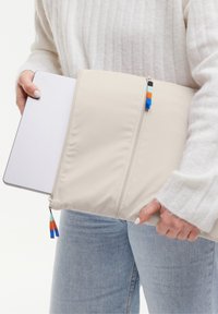 Custodia per laptop beige con due cerniere, texture in tessuto e tirazip colorati in blu, arancione e teal, tenuta da una persona in un maglione chiaro.