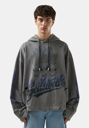 Giovane uomo che indossa una felpa grigia oversize con grafica di teschio e fiamme e dettagli logori, abbinata a jeans blu chiaro, sfondo neutro.