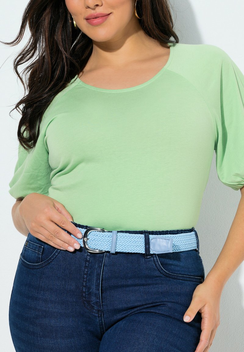 Frau trägt ein hellgrünes kurzärmeliges Oberteil, das in eine dunkelblaue Jeans mit einem hellblauen gewebten Gürtel und silberner Schnalle gesteckt ist.