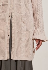 Světle růžový pletený cardigan s kabelovými vzory, žebrovanými akcenty a dvěma velkými kapsami. Má zapínání na knoflíky a zvonové rukávy.