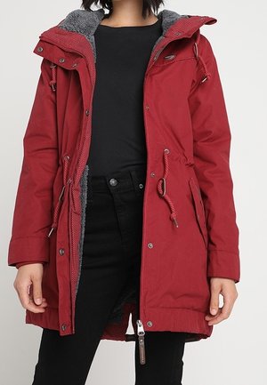 Personne portant un parka rouge à capuche avec une doublure polaire grise sur un haut noir et un pantalon noir, debout devant un fond uni.