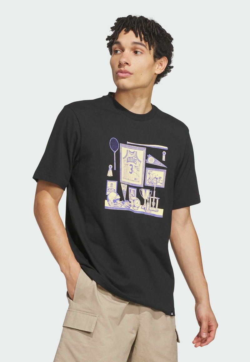 Jeune homme portant un t-shirt noir avec des motifs de souvenirs sportifs et un short cargo beige, regardant vers sa gauche avec une main dans la poche.