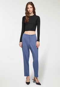 Giovane donna con capelli castani ondulati che indossa un top nero a maniche lunghe corto, pantaloni blu a vita alta e décolleté neri a punta.