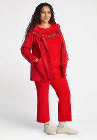 Röd vattentät jacka med huva, kombinerad med en matchande röd T-shirt och byxor. Krämfärgad sneakers fulländar outfiten. Slät textur, avslappnad passform.