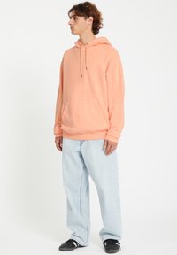 Haut à capuche pêche avec une poche avant, cordon de serrage à la capuche et poignets côtelés, associé à un jean bleu clair à larges jambes et des baskets noires.