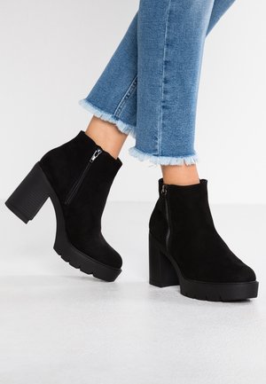 Zwarte suède platform enkelaarsen met blokhakken en ritsen aan de zijkant, gedragen met blauwe jeans met een rafelige zoom op een witte vloer.