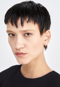 Close-up of a person with short, straight black hair, wearing a light earring and a simple dark top, featuring natural skin texture and subtle freckles.