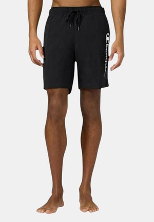 Pantaloncini neri a metà coscia con vita con coulisse e logo verticale bianco sulla gamba destra, indossati da una persona in piedi a piedi nudi.