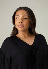Blusa nera plissettata con maniche ampie, scollatura a V e dettaglio con nodo sulla parte anteriore. Tessuto testurizzato con accenti di cucitura sottili.