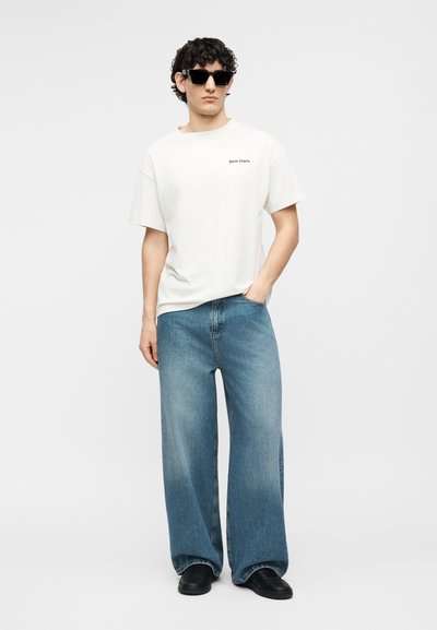 Άνδρας που φοράει μαύρα γυαλιά ηλίου, λευκό κοντομάνικο μπλουζάκι με μικρό κείμενο, φαρδιά μπλε τζιν και μαύρα παπούτσια, στέκεται με το ένα χέρι στην τσέπη.