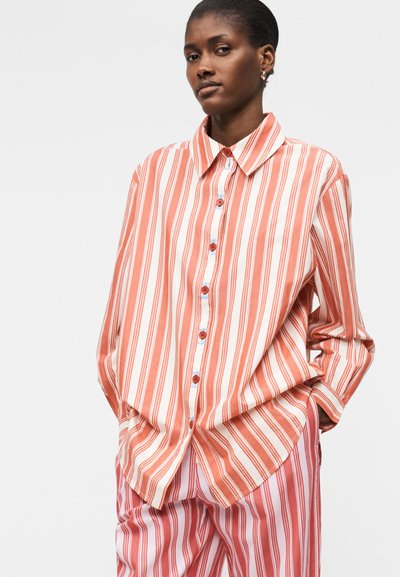 Γυναίκα που φοράει άνετο πουκάμισο με μακριά μανίκια με πορτοκαλί και λευκές κάθετες ρίγες και αντίστοιχο ριγέ παντελόνι, τα χέρια στις τσέπες.