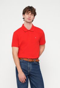 Piros, sima pamutból készült galléros póló, bal mellrészen hímzett logóval; kék farmernadrággal és barna övvel viselve.