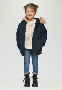 Marineblauwe pufferjack met een nepbontkap, romige gebreide trui, lichtblauwe jeans en zwarte veterschoenen. Bevat knopen en zakken.