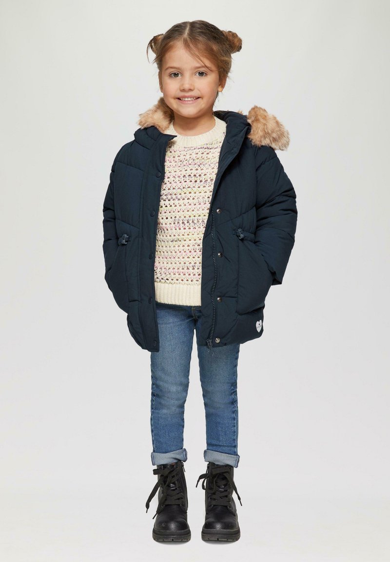 Marineblauwe pufferjack met een nepbontkap, romige gebreide trui, lichtblauwe jeans en zwarte veterschoenen. Bevat knopen en zakken.