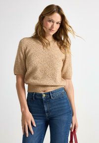 Maglione beige a maglia con maniche corte, caratterizzato da un motivo a giorno, abbinato a jeans blu a vita alta e una borsa rossa.