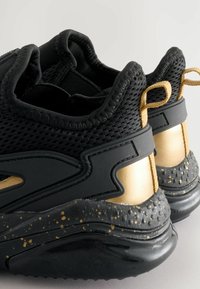 Baskets noires de sport avec une tige en mesh respirant, accents dorés, semelle texturée avec des éclats noirs et dorés, et languettes au niveau du talon.