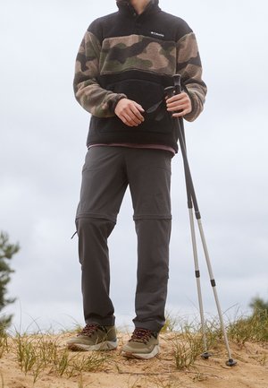 Personne portant une veste polaire camouflage et un pantalon sombre, tenant des bâtons de randonnée, debout sur un sol sablonneux avec de l'herbe sous un ciel nuageux.