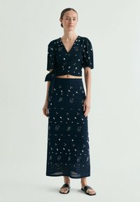 Mujer vestida con un conjunto de dos piezas azul marino con patrones blancos de cachemira, top cruzado corto, falda a juego hasta el tobillo y sandalias negras.