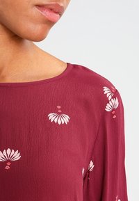 Blouse bordeaux à texture lisse, ornée de broderies florales blanches et roses, col rond et manches longues.