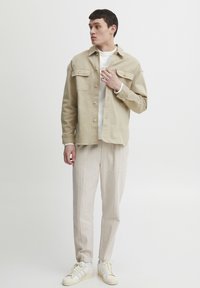 Beige denim jacket with two chest pockets, layered over a white sweatshirt, paired with light linen trousers and white sneakers.