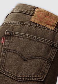 Jeans de mezclilla marrones que cuentan con un bolsillo trasero con costuras en contraste y un parche de cuero texturizado en color marrón claro que muestra la información de la marca.